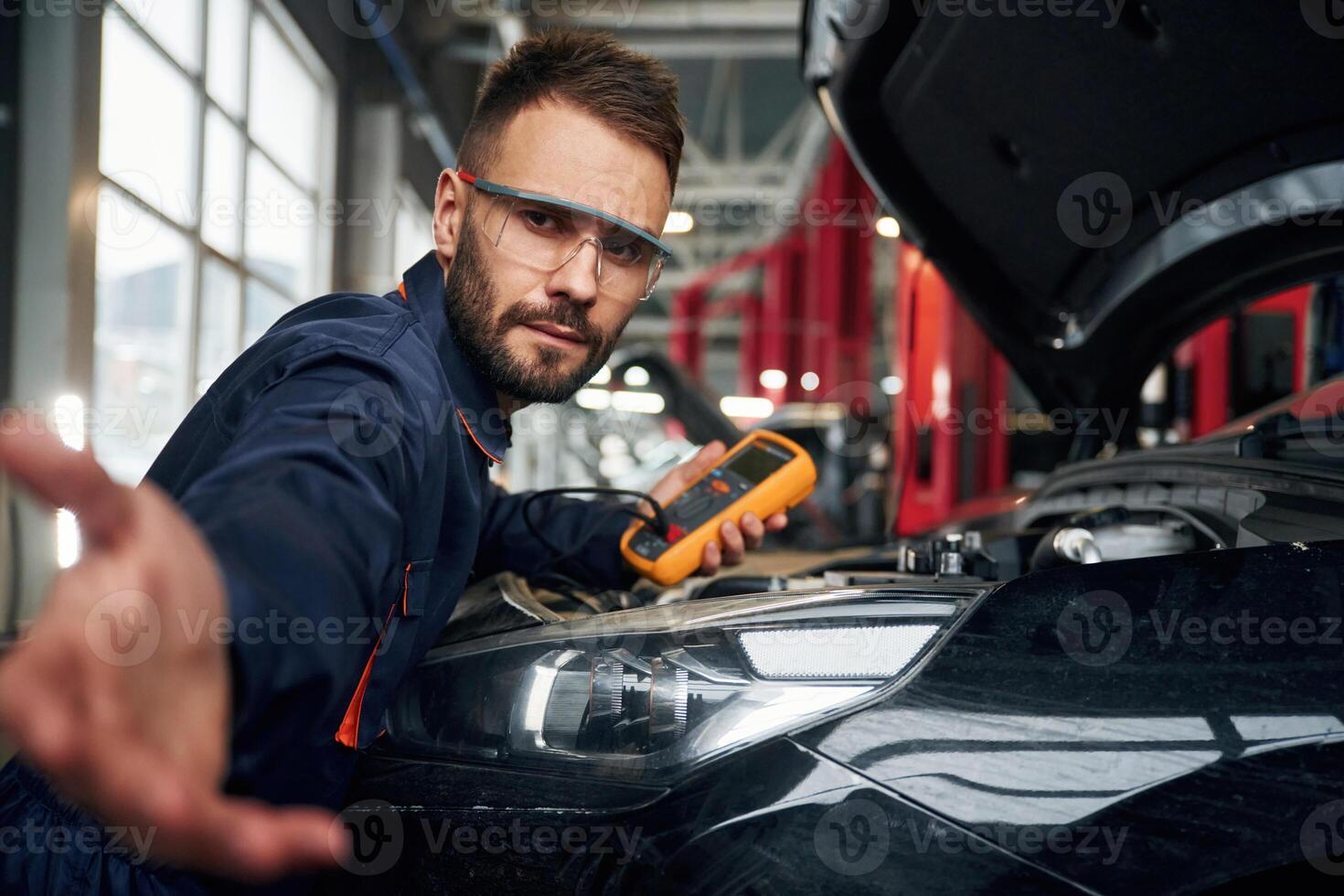 Yellow colored ,device for measuring electricity is in hands. Car repairman is in the garage with automobile photo