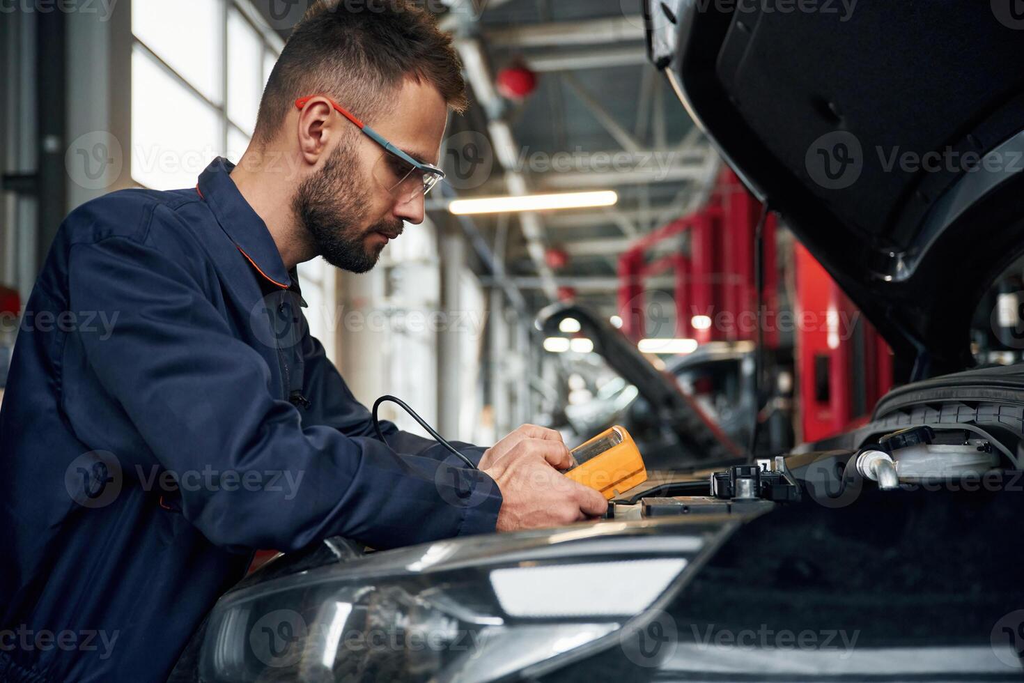 Yellow ,colored device for measuring electricity is in hands. Car repairman is in the garage with automobile photo