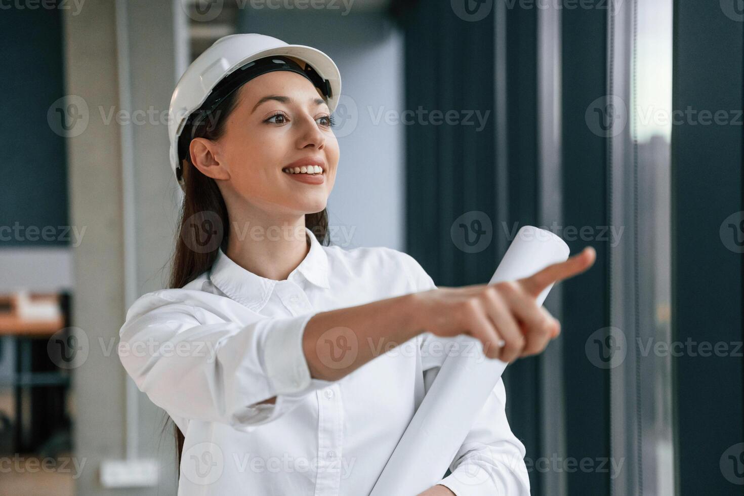 Pointing, by finger to, the side. Female architect in hard hat and in white clothes is indoors photo