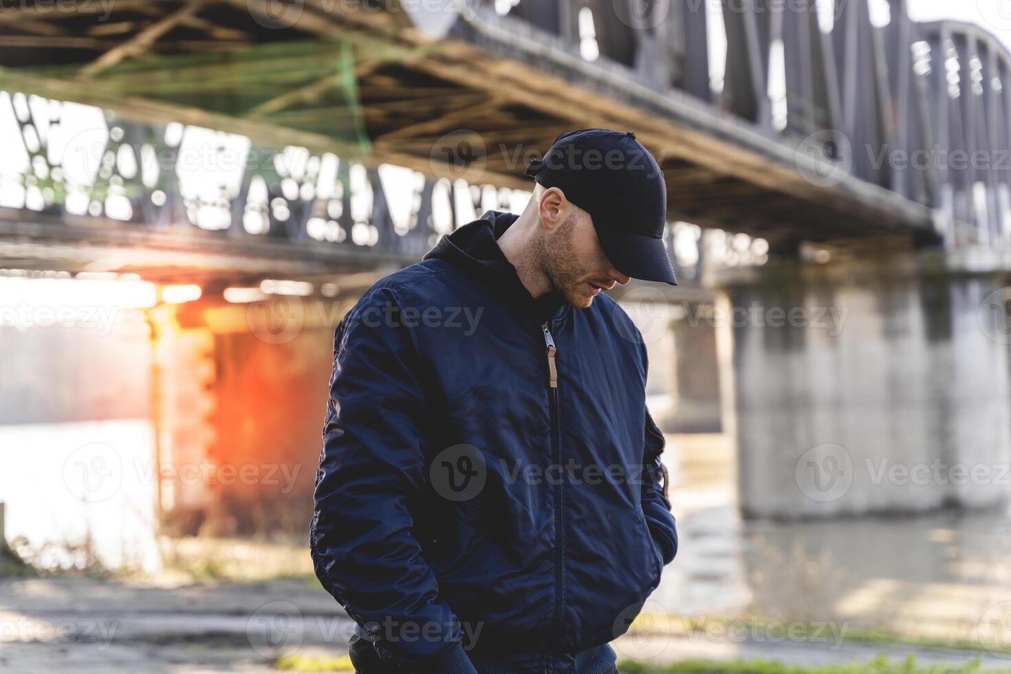 a young rapper in winter clothing on the banks of a river under a bridge photo
