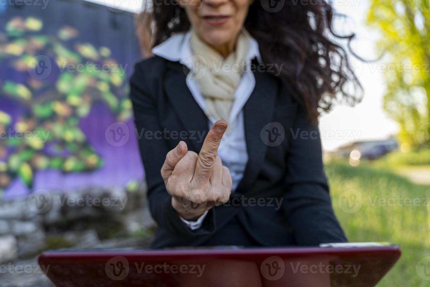 Beautiful middle-aged manager wearing red glasses shows middle finger gesture in displeasure while working on her laptop outdoors photo