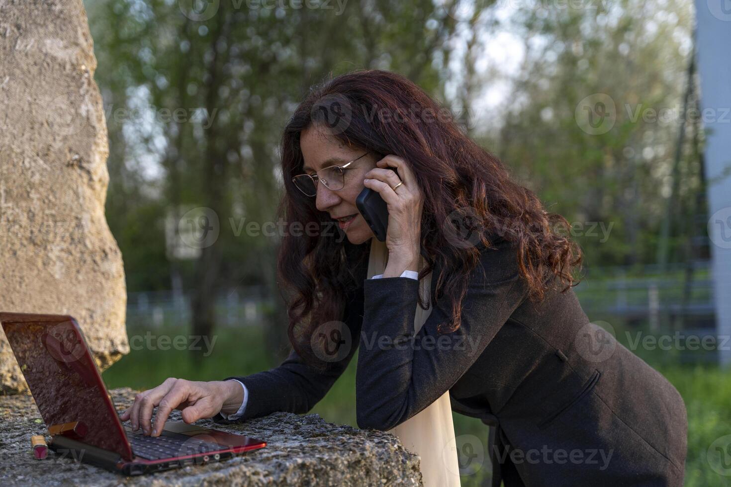 beautiful middle-aged businesswoman talking on cell phone and working on laptop leaning on concrete cube as desk outdoors photo