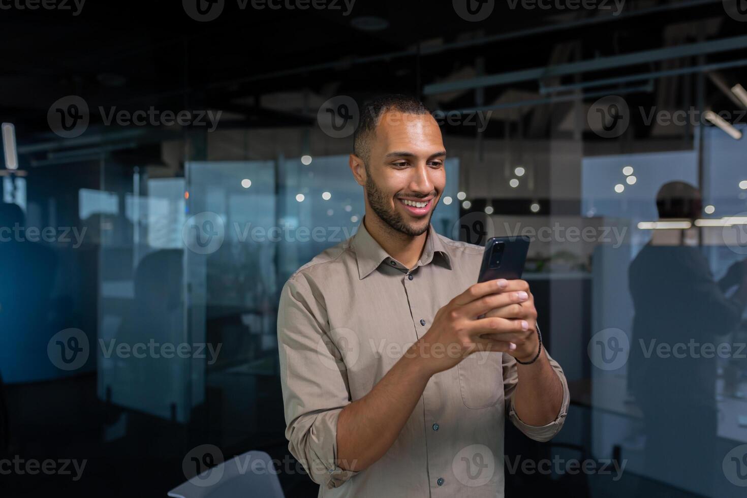 Smiling african american businessman inside office in shirt using phone, man typing message and browsing internet pages online standing near window at workplace in the evening. photo
