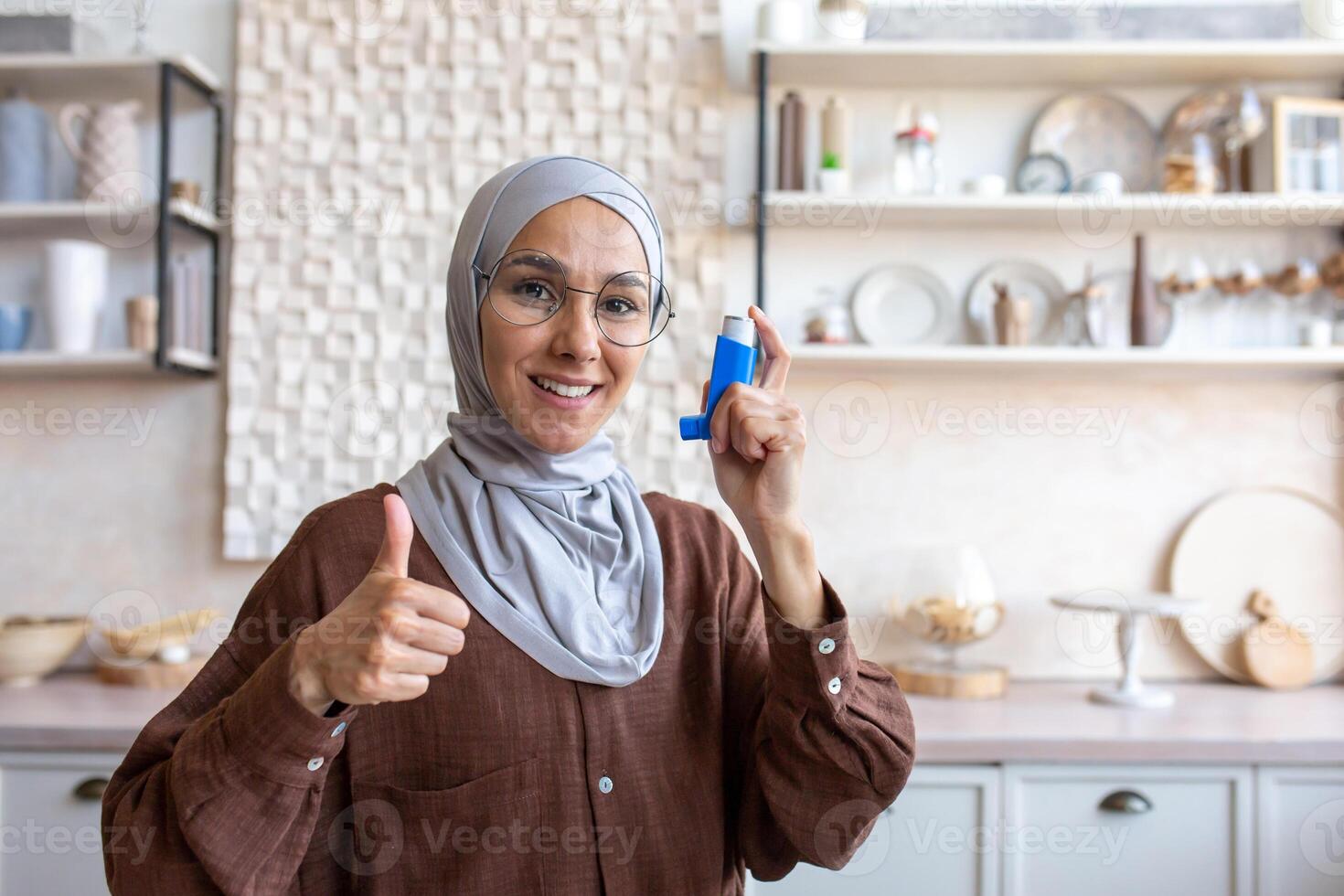 Close-up photo portrait of a smiling young Muslim woman in a hijab sitting at home holding an asthma spray in her hand and pointing super finger at the camera.