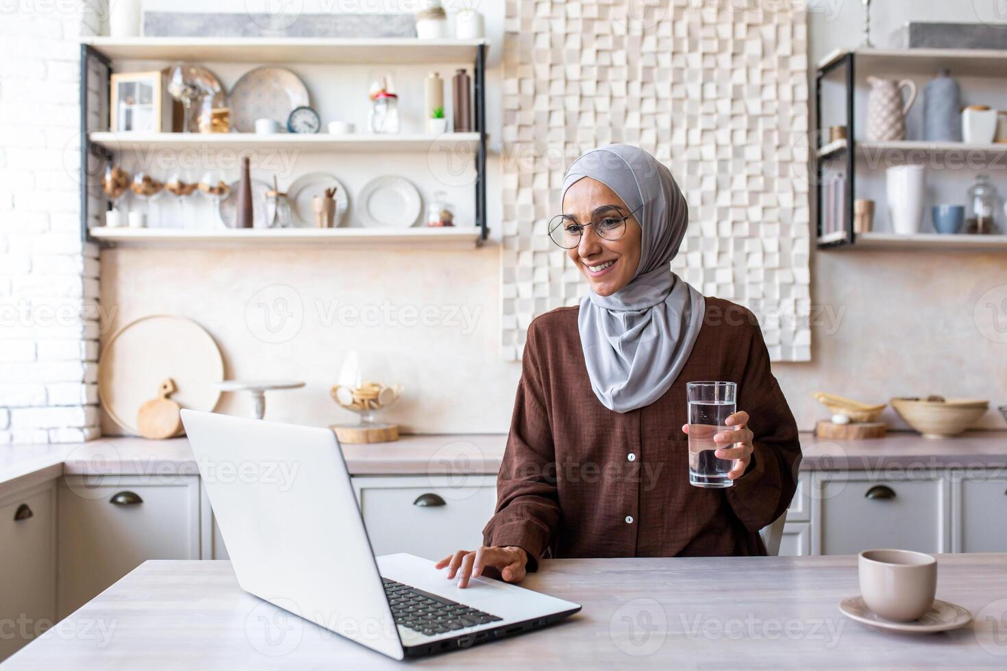 Smiling young Muslim woman in hijab working and studying at home using laptop. Sitting in the kitchen at the table and holding a glass of clean drinking water. photo
