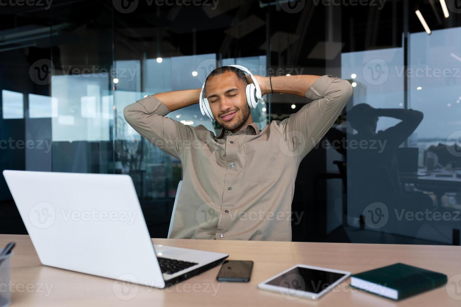Successful young african american businessman relaxing in office, man in shirt and headphones listening to music online radio and podcasts, sitting at desk using laptop at work. photo