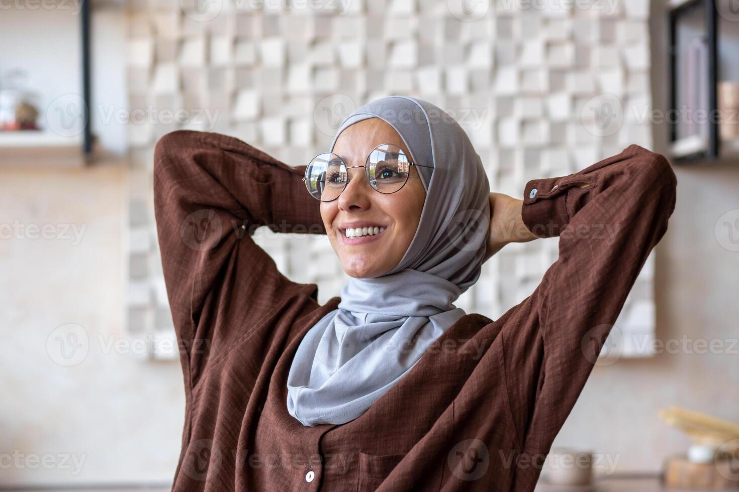 Close-up photo. Young muslim woman freelancer in hijab sitting at table at home after working day using laptop. She threw her hands behind her head, resting, smiling, satisfied with the result. photo