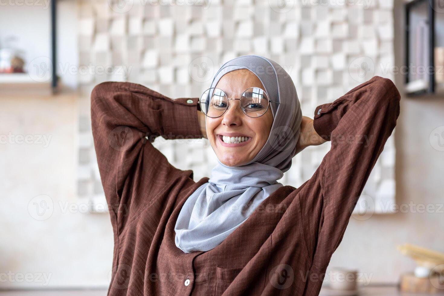 Close-up photo portrait of a young Muslim woman in a hijab with her hands behind her head and resting at home, looking at the camera smiling and satisfied.