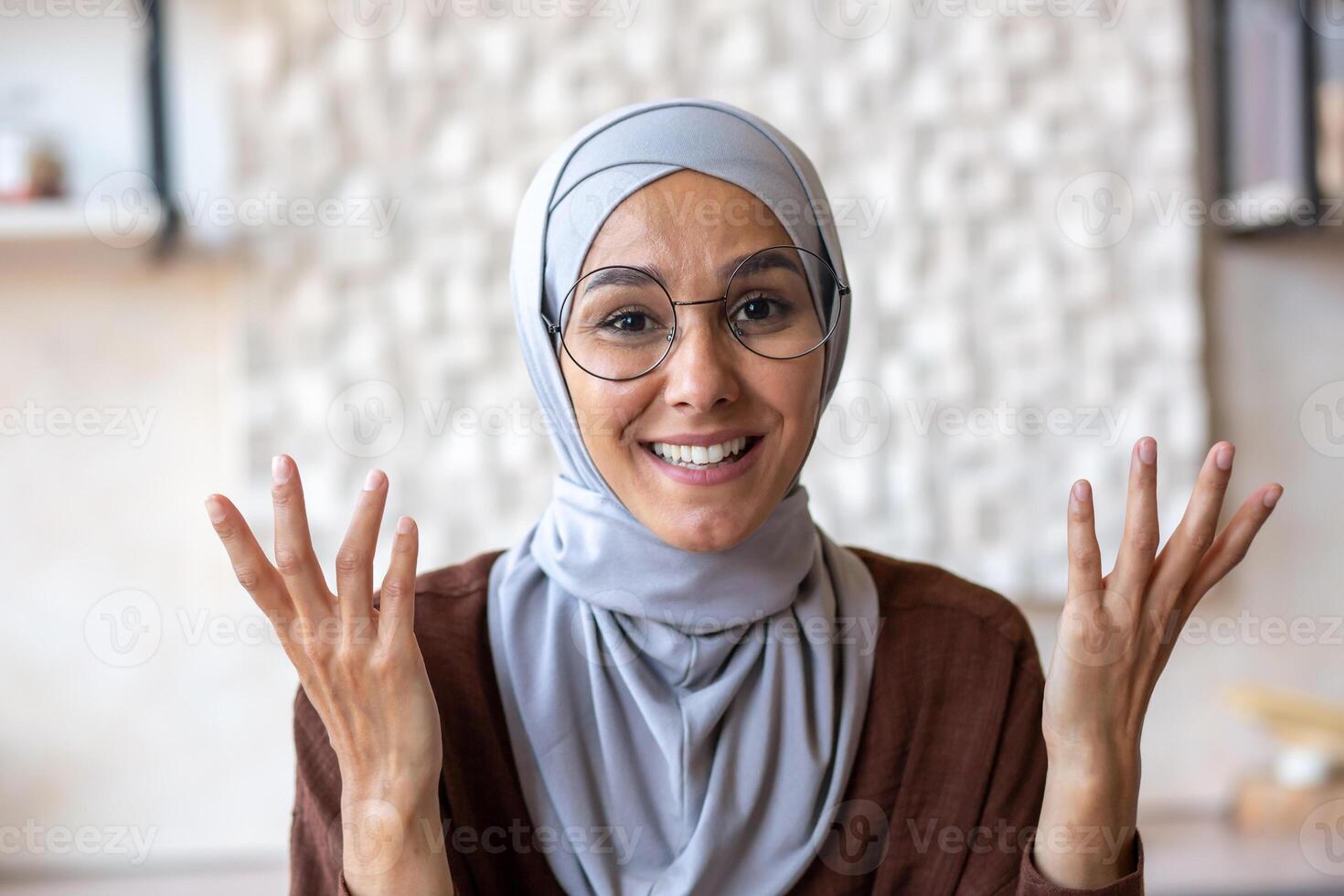 Close-up photo. Arab young woman in hijab and glasses talking to camera from home. Gesturing with hands, smiling. photo
