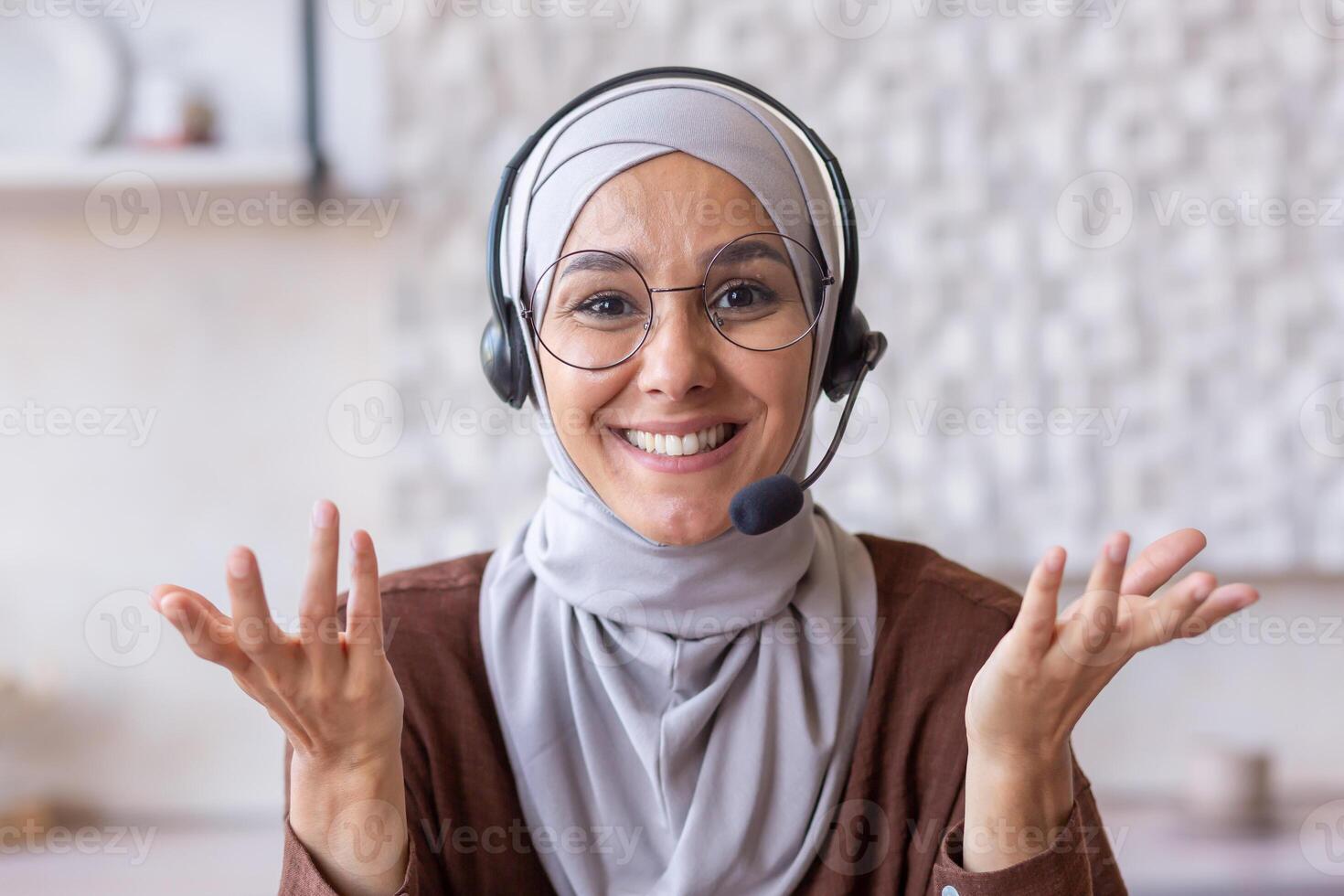 Hijab woman working remotely at home, Muslim woman with call headset smiling and looking at camera, talking to client online in kitchen, webcam view pov. photo