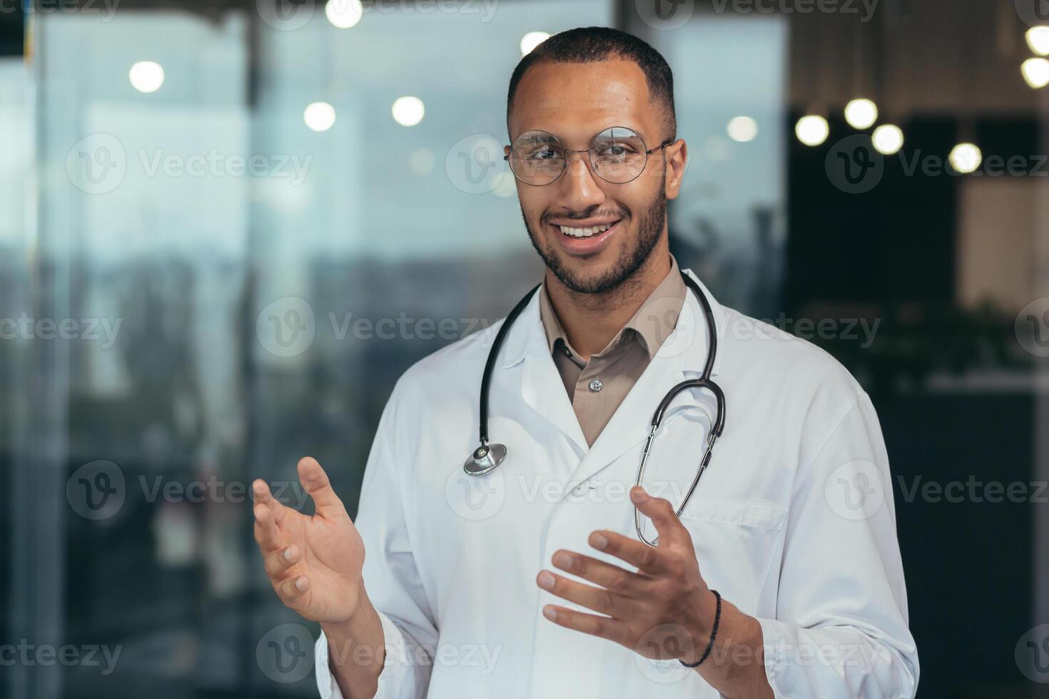 Close-up photo of hispanic doctor smiling and looking at web camera talking to colleagues and patient on call, man in medical coat and stethoscope working inside clinic office.