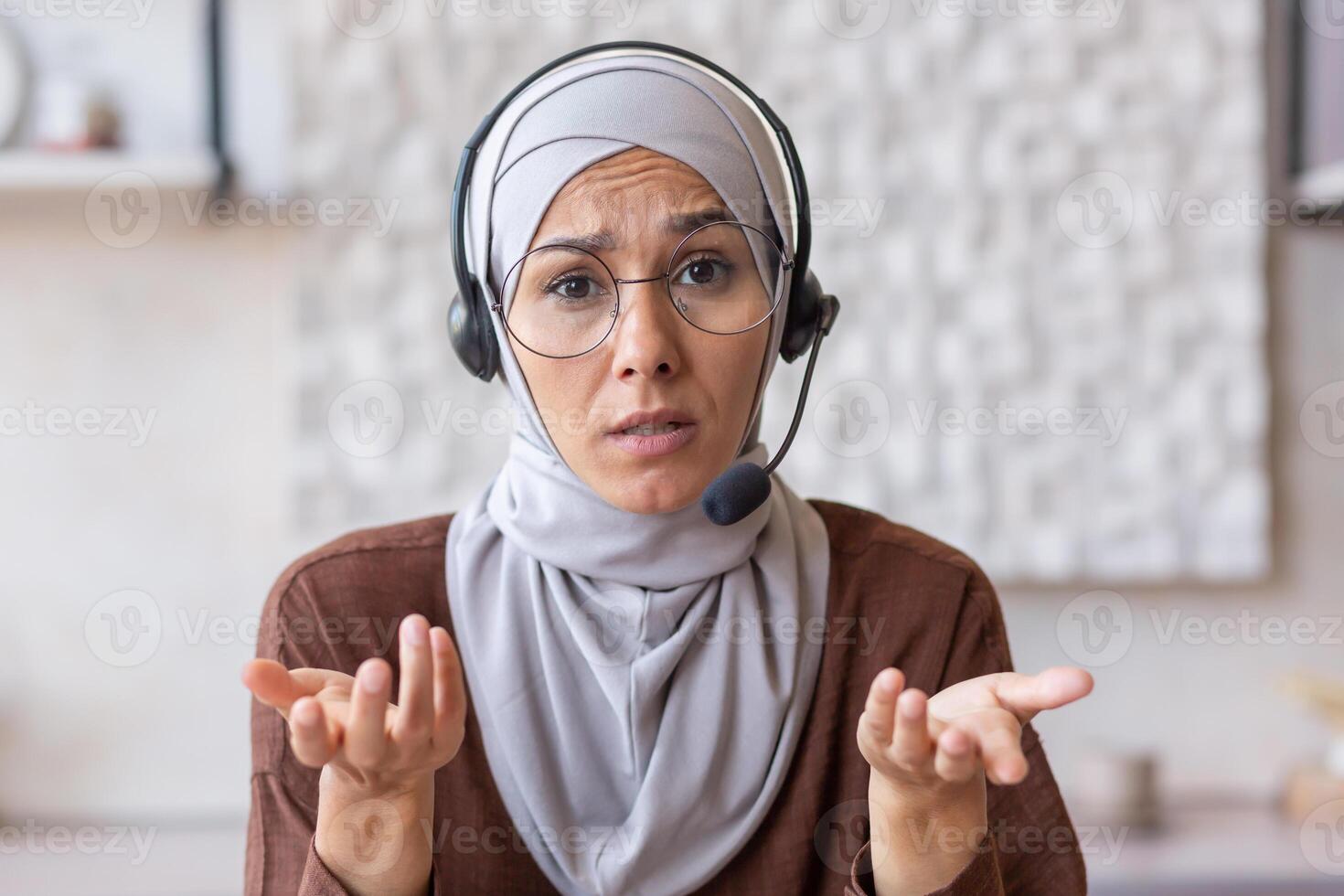 Close-up photo. Serious and frustrated young muslim woman in hijab and wearing headset and glasses talking and explaining on call to camera at home. photo