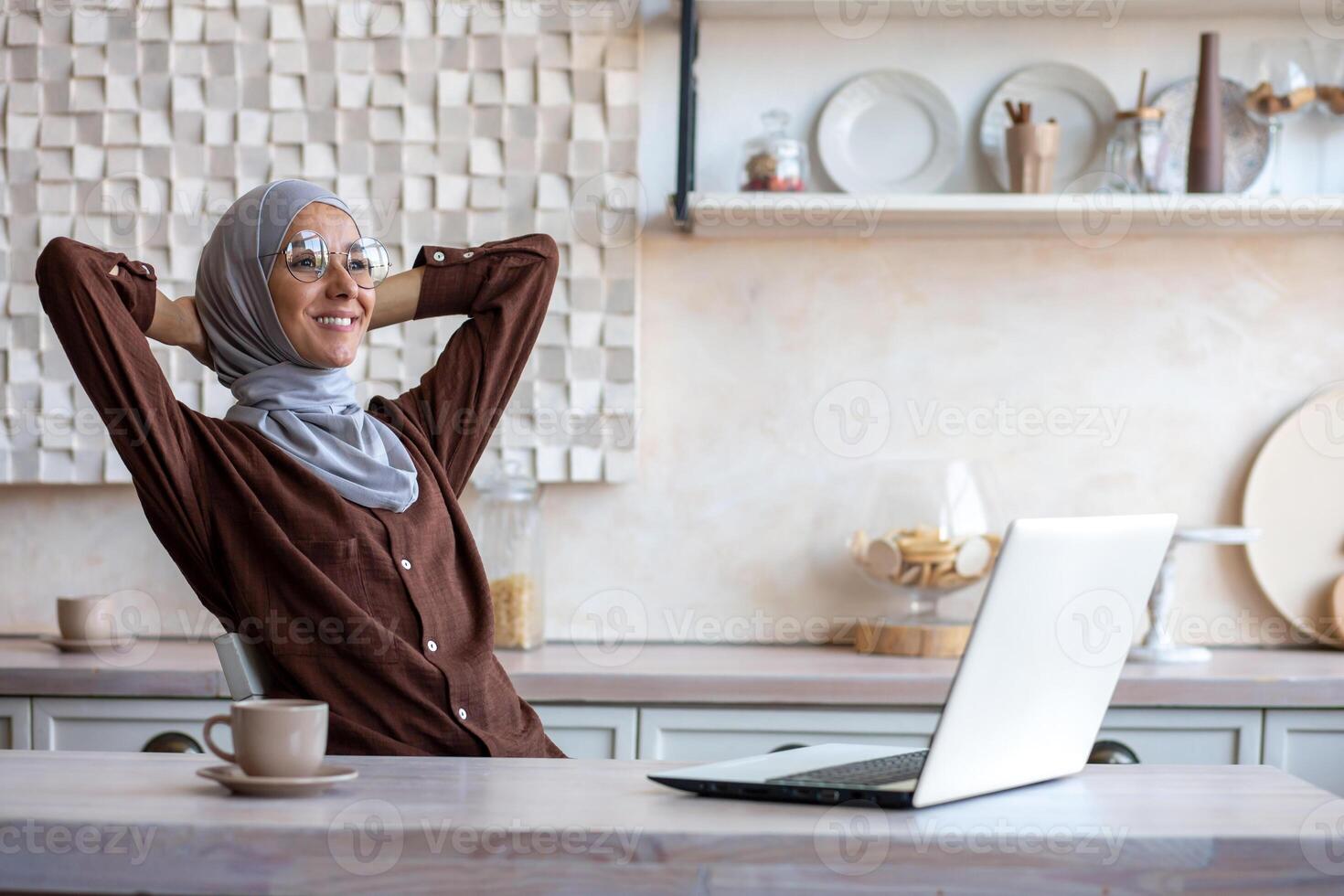 Young muslim woman freelancer in hijab sitting at table at home after working day using laptop. She threw her hands behind her head, resting, smiling, satisfied with the result. photo
