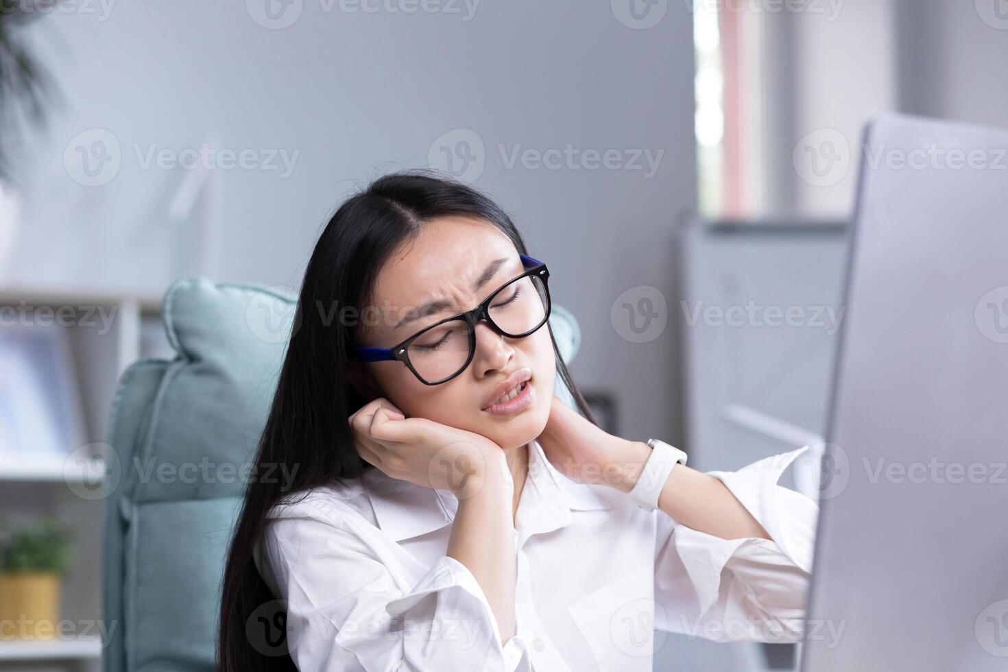 Close up photo, youngAsian woman office worker, freelancer, business woman in glasses sitting at the table at the computer, tired, exhausted,doing neck massage with two hands, taking a break from work photo