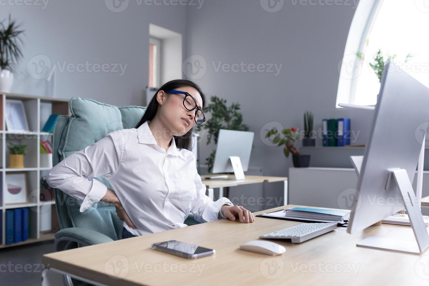 Young beautiful azitaka businesswoman wearing glasses feeling exhausted at work. Holds his back with two hands, massages, feels pain and fatigue. Sitting at the desk at the computer, needs a break. photo