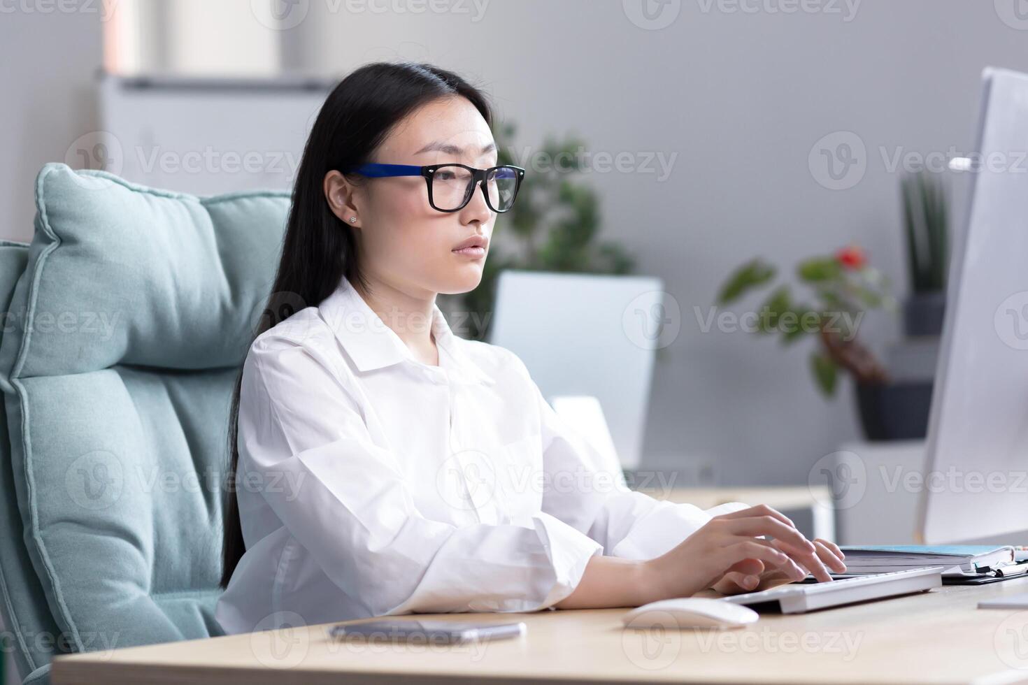 Young Asian girl student in glasses sits and studies at the computer online remotely at home at the table, typing on the keyboard, passing the exam, serious. photo