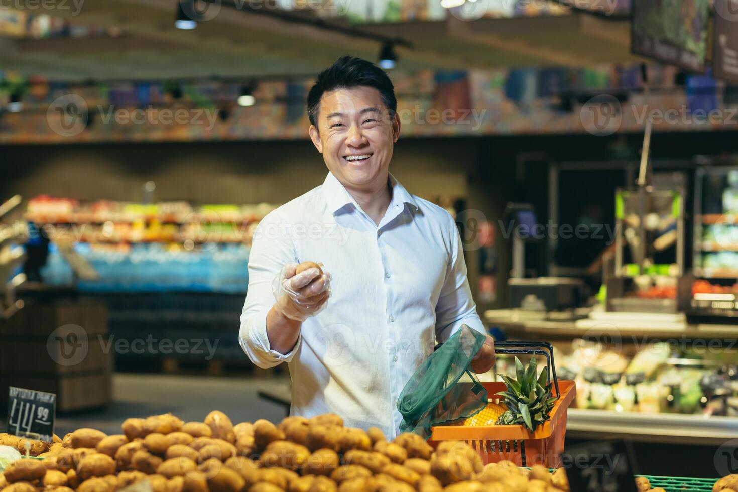 A young handsome Asian man, businessman is standing in a supermarket in the vegetable section, holding a basket of fruits and potatoes in his hands. He looks at the camera, smiles. photo