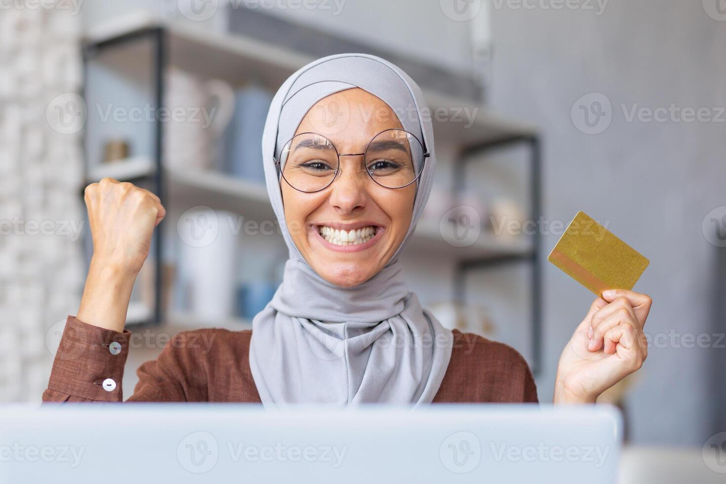 Close-up photo. Muslim young woman in hijab sitting at home using laptop and holding credit card. Happy on camera. Made a cherished dream come true, made an online purchase. photo