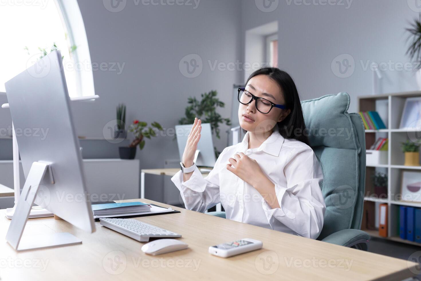 Heat in the workplace. Young Asian woman sitting in the office at the desk waving her hand, it's very hot, she feels bad, she needs fresh air, take a break. photo