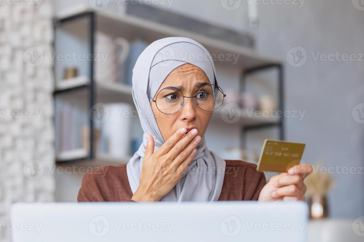 Close-up photo. Portrait of a young Muslim woman in a hijab sitting at home using a laptop and holding a credit card in her hand. He looks at her in shock, covers his mouth with his hand. photo
