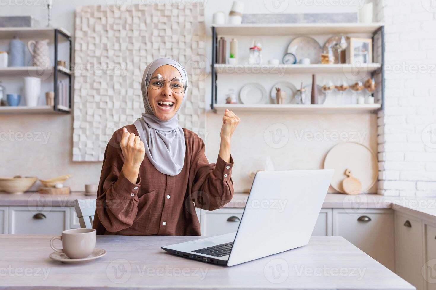 Young happy Muslim student woman in hijab sitting at home, studying on laptop. Shows a yes gesture with his hands to the camera. Passed an online exam, a letter from the university, good results. photo