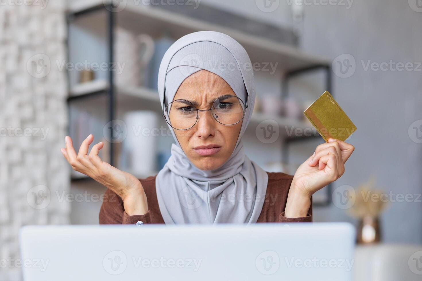 Close-up photo. Credit card problems. Worried young Muslim woman in hijab sitting in front of laptop. He looks at the monitor in shock, spreads his hands. photo