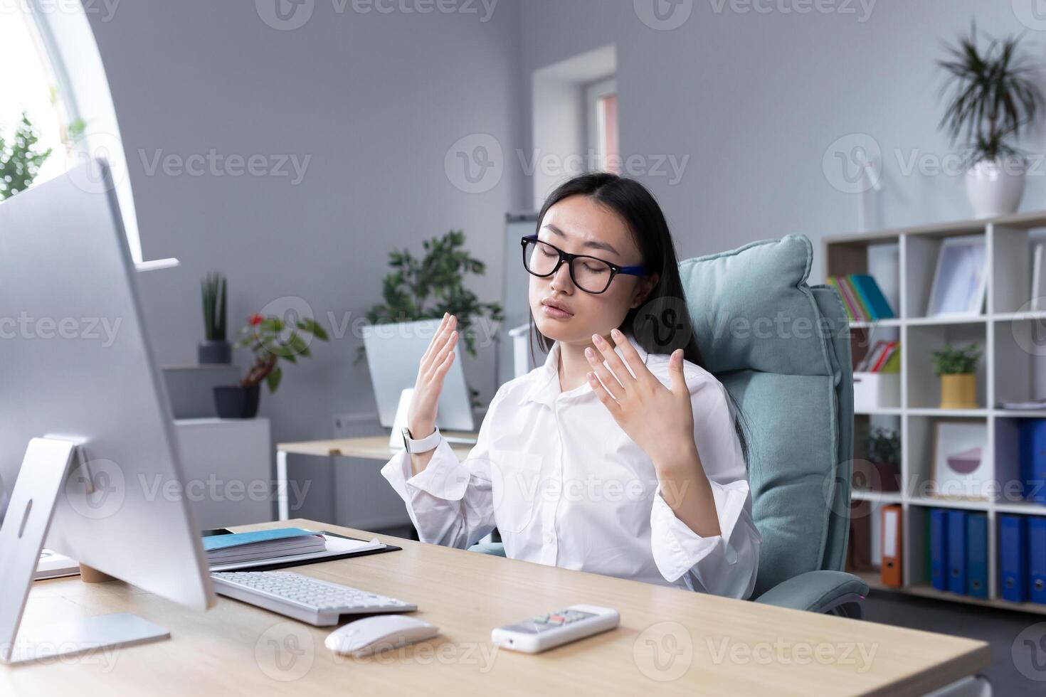 Heat in the workplace. A young Asian woman sits in the office at the desk, waves both hands at her face, it is very hot, she feels bad, she needs fresh air, take a break. photo