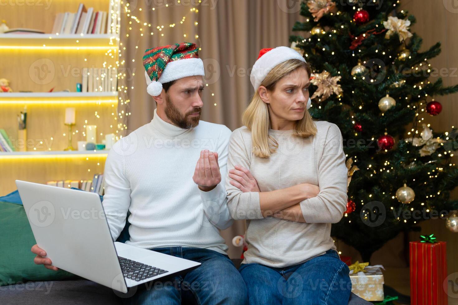 A stingy man does not allow a woman at home to make purchases in the online store online shopig, a married couple, a man and a woman, are sitting on the sofa at home near the Christmas tree in New Year's hats, on New Year's holidays. photo