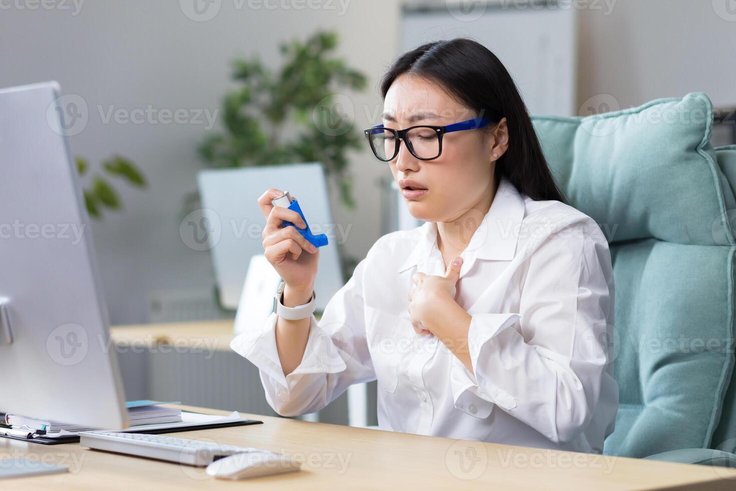 Allergy, feeling unwell at the workplace. A young Asian woman in glasses coughs, chokes, uses an inhaler, sneezes in her throat, has allergies, asthma. He is sitting at his desk in the office. photo