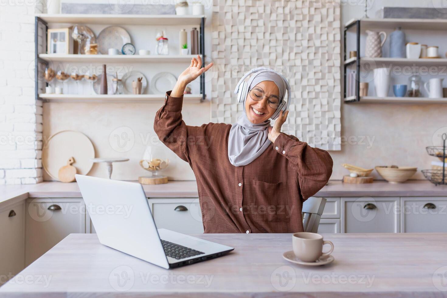 Young arab woman businesswoman, hijabi freelancer working from home online at home using laptop. She sits with headphones, listens to music, raised her hands, dances, rests, takes a break. photo