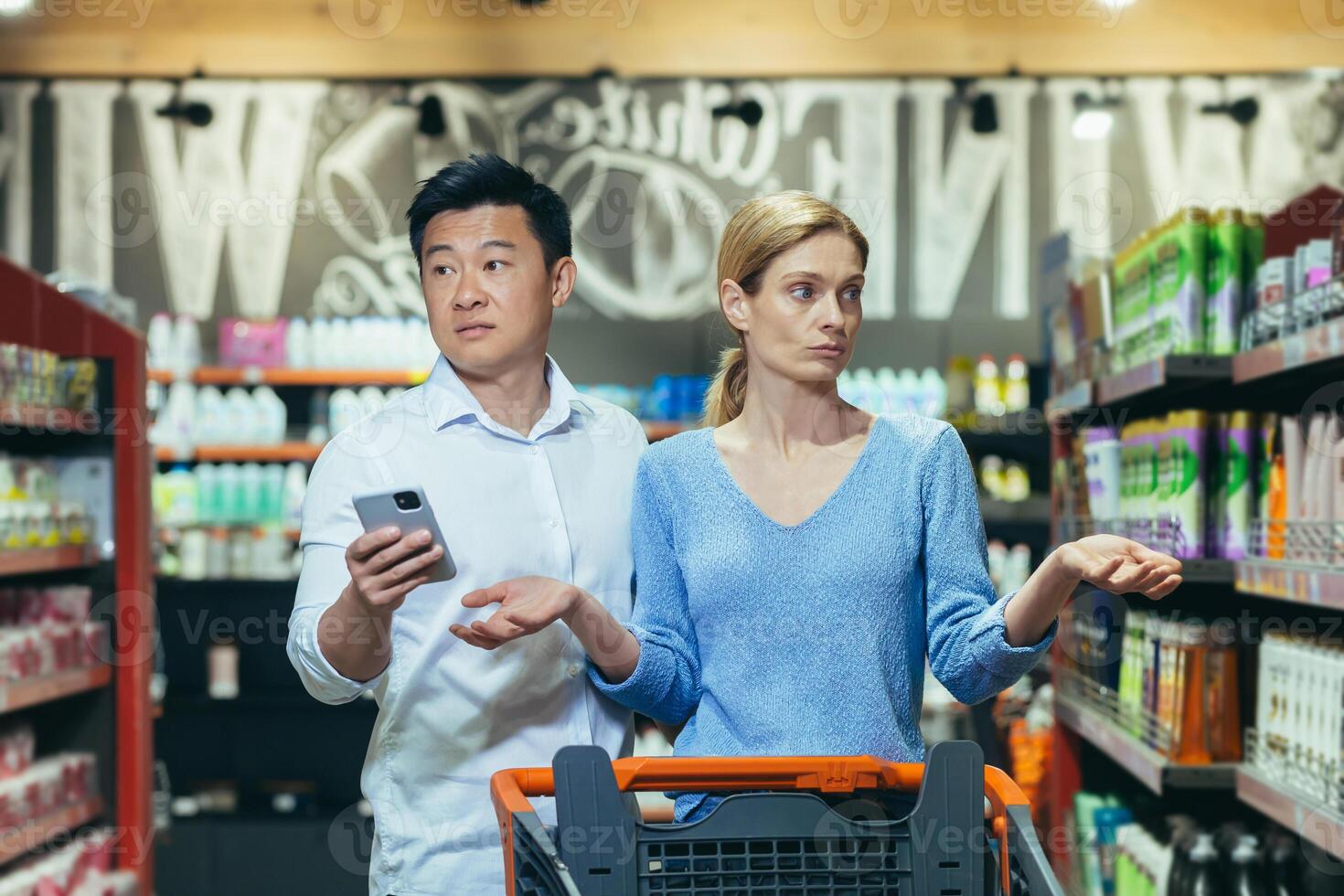 A couple of buyers, a man and a woman, confused in the supermarket, cannot choose the necessary product, an Asian man with a phone photo
