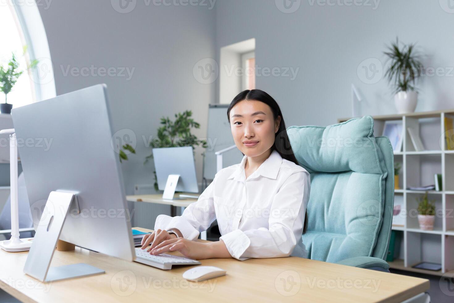 Beautiful Asian woman works at the computer in the office, looks at the camera and smiles, business woman in a white shirt typing on the keyboard. photo