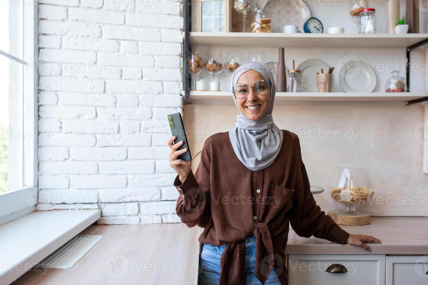 A young Muslim woman in a hijab is standing at home in the kitchen by the window and using the phone. Smiling, she looks at the camera. photo