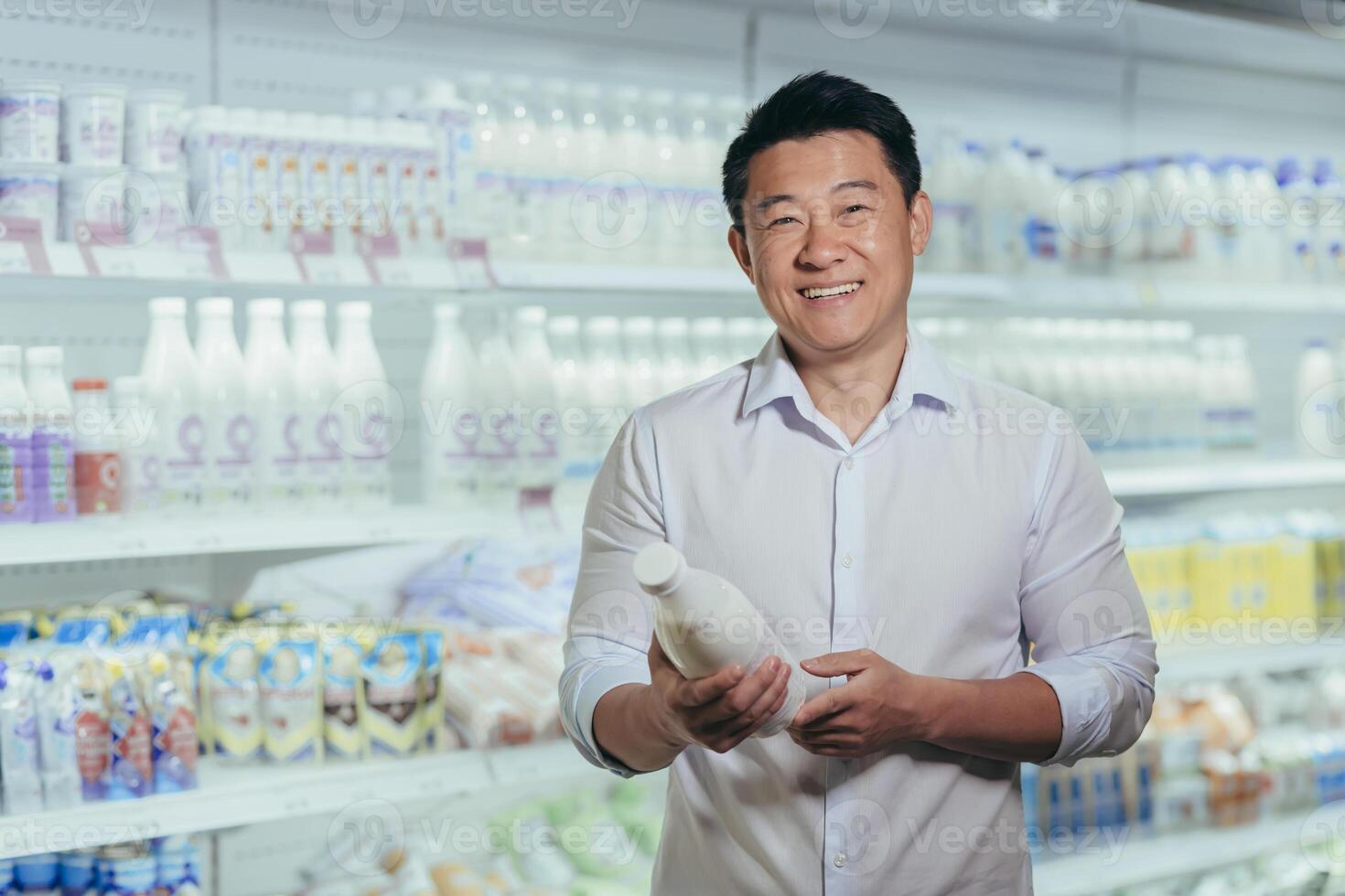 Portrait of happy and satisfied asian shopper in supermarket in dairy department, man holding bottle of milk smiling and looking at camera near refrigerators photo