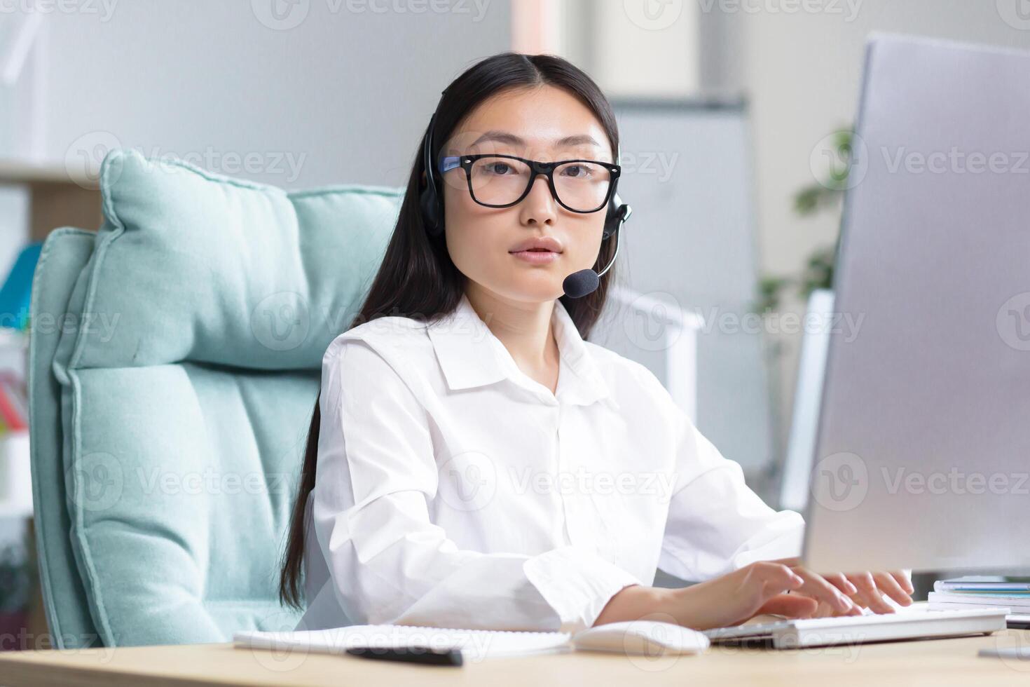 Portrait of a young Asian woman in headphones with a microphone. Works as a consultant in a call center. Sitting at the table in glasses at the computer, looking at the camera, smiling. photo