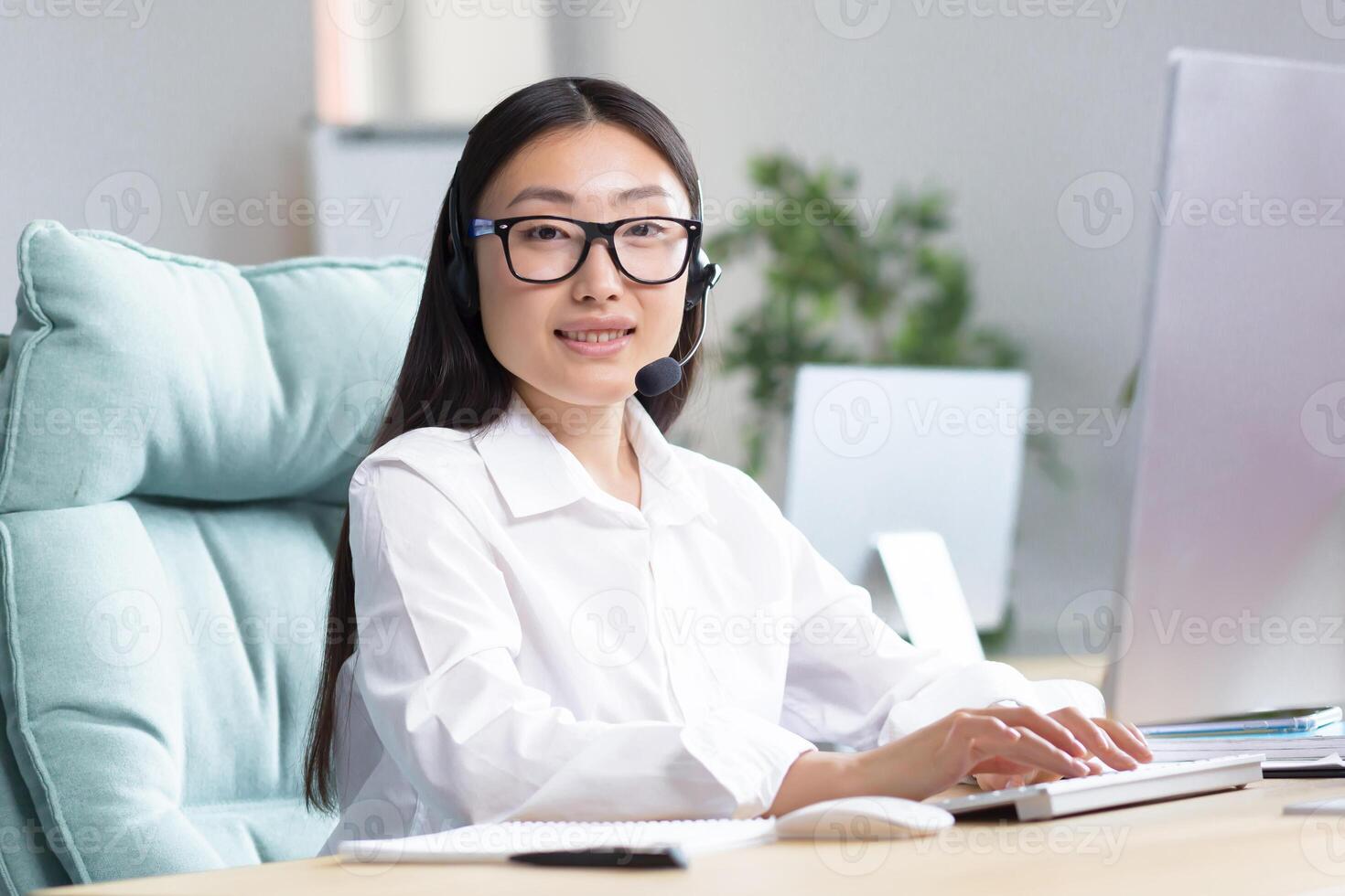 Portrait of a young beautiful Asian woman in headphones with a microphone. Works as a consultant in a call center, answers calls. Sitting at the table, looking at the camera, smiling. photo