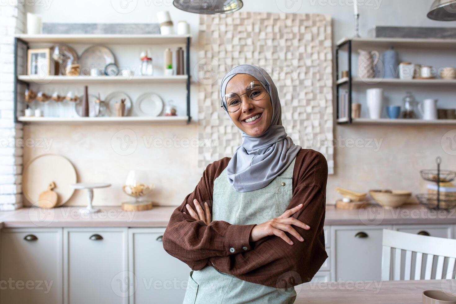 Portrait of young Muslim woman food blogger, coach, chef in hijab standing at home in kitchen in apron, arms crossed on chest, posing, smiling and looking at camera. Conducts online courses, webinars. photo