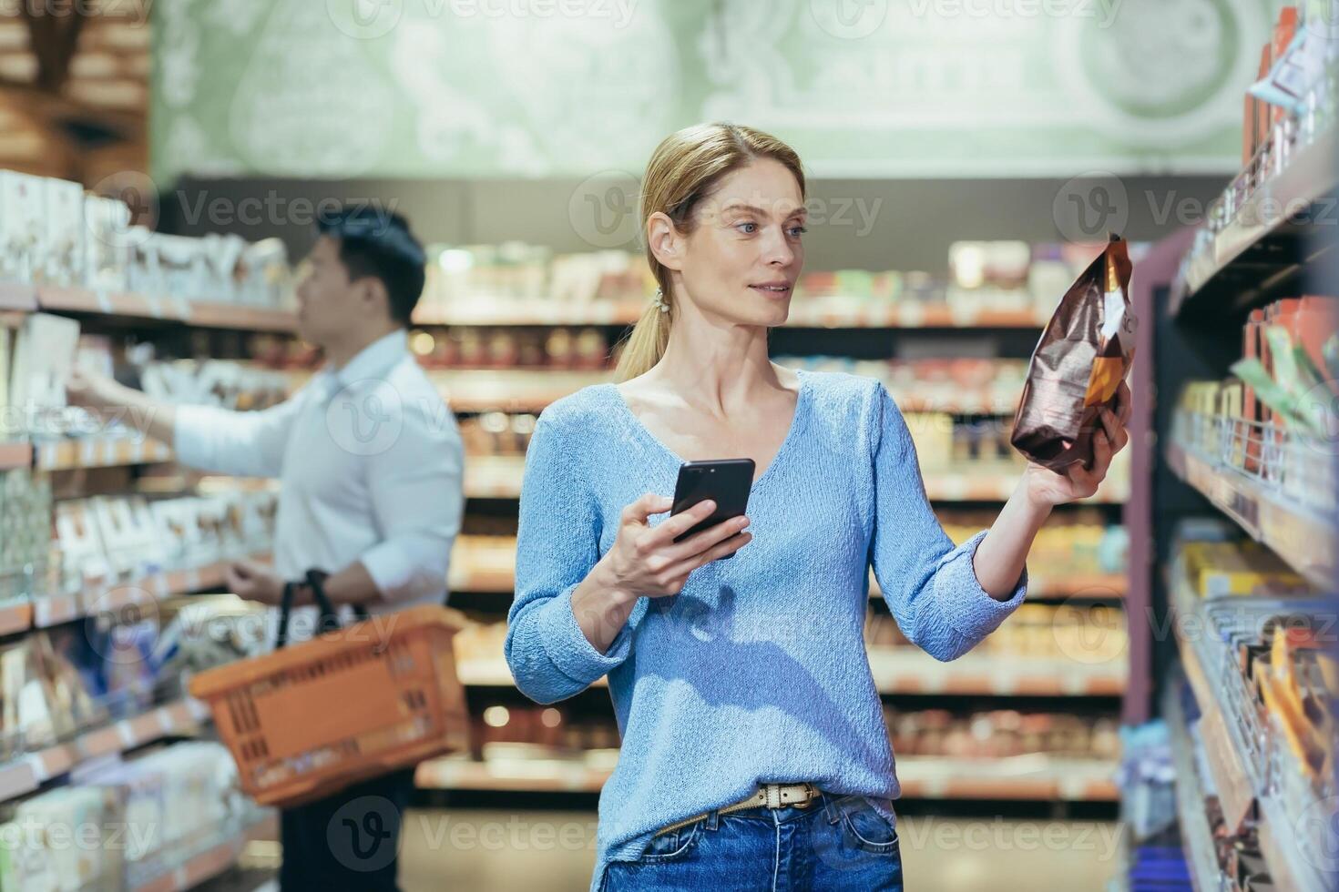 A beautiful young blonde woman is standing in a supermarket. She is holding a phone and a packet of breakfast cereal. Chooses, buys, reads the label. Shoppers in the background. photo