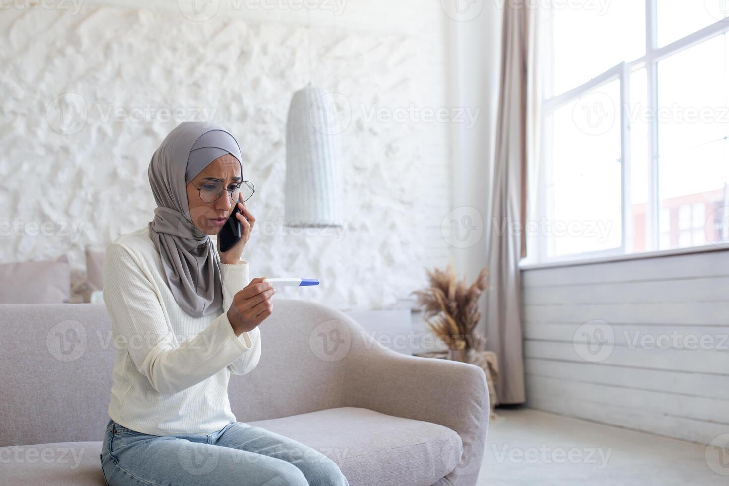 Upset and disappointed young arab woman in hijab sitting on sofa at home and talking on the phone. Holds a quick and one-time pregnancy test with a negative result. photo
