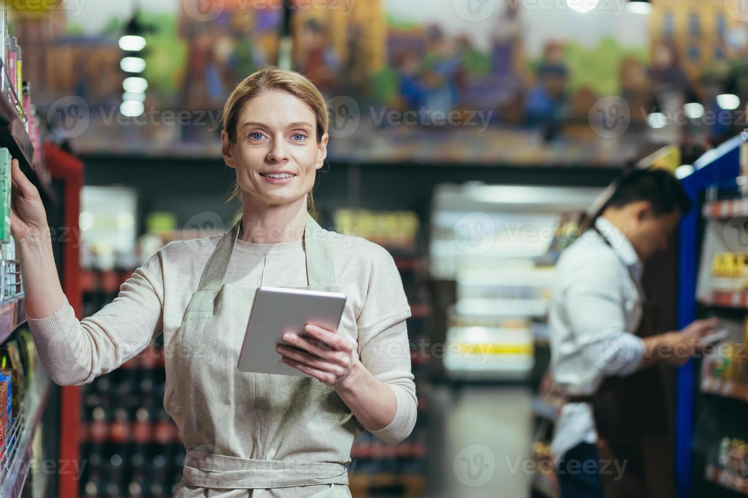 Portrait of an experienced female seller in a supermarket, the manager is looking at the camera and smiling, holding a tablet computer in her hands, the saleswoman is among the shelves with goods in the store photo