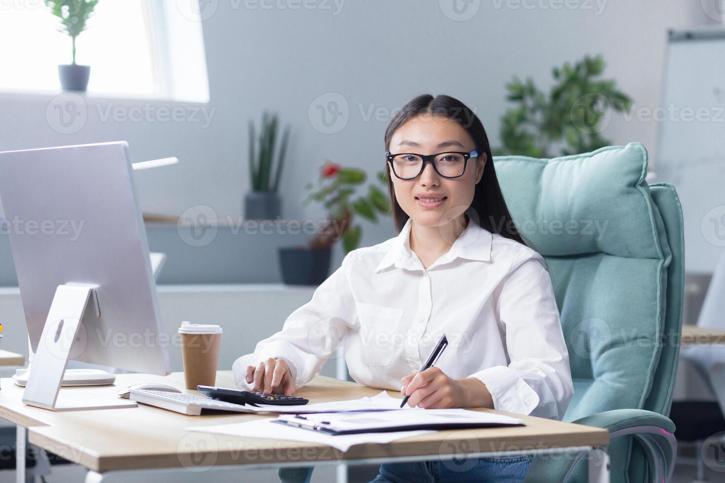 Workaholic. A young Asian business woman, sitting at the table drinking coffee to go, a paper cup of coffee is standing on the desktop near the computer. He looks at the camera, smiles. photo