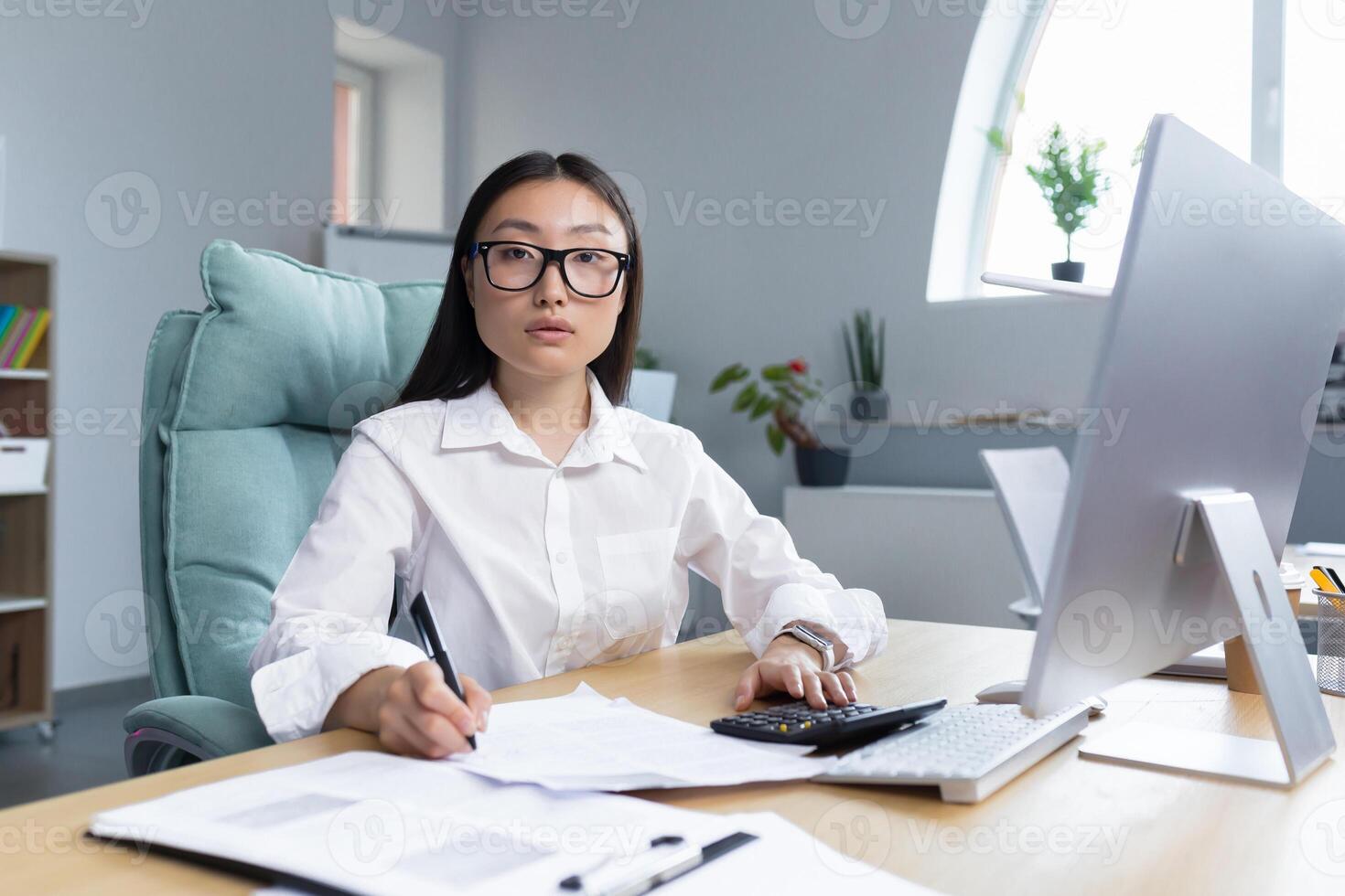 Work with documents. Portrait of a young beautiful business woman Asian accountant works with documents and reports. Sitting at the desk in the office, writing.He looks at the camera. photo
