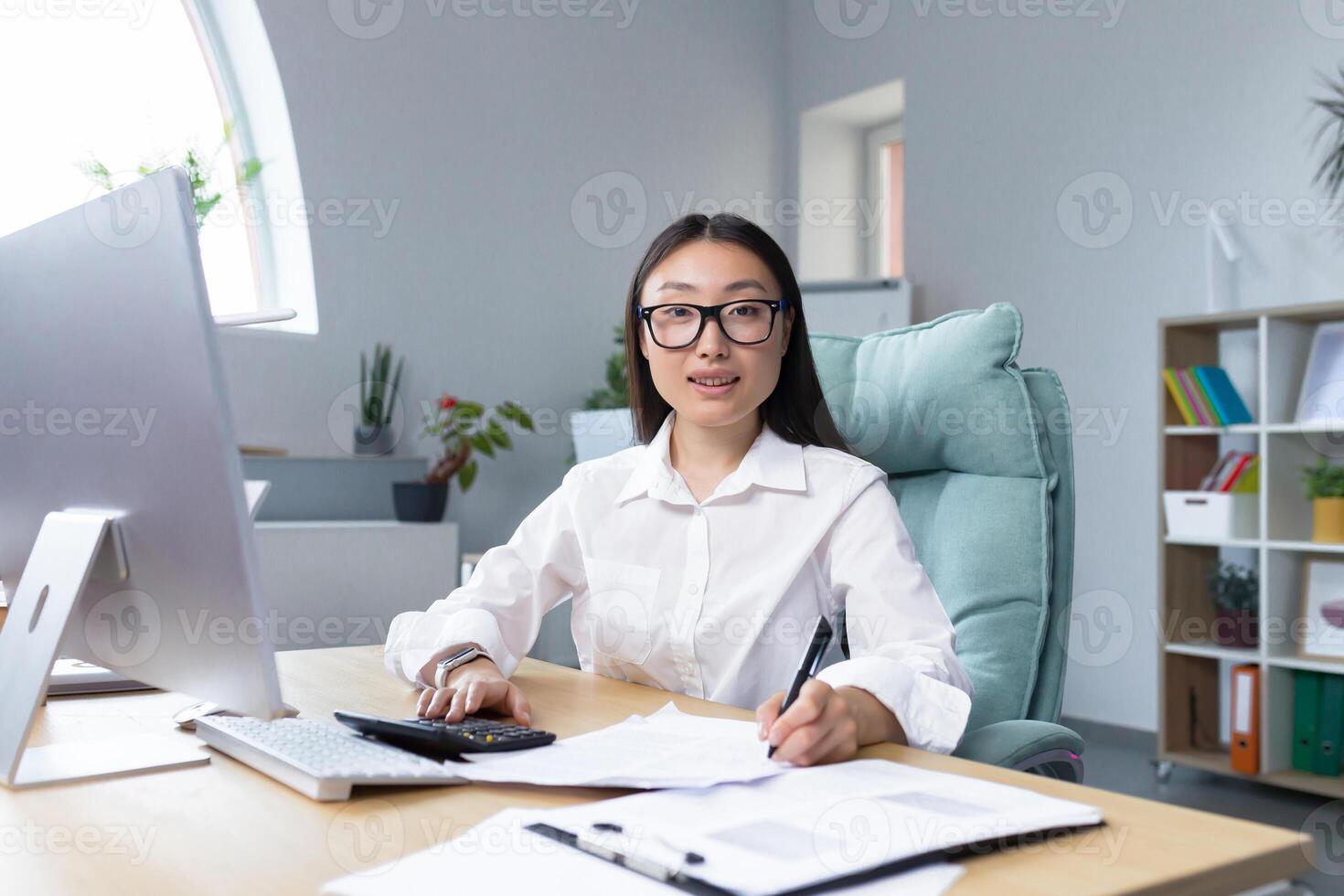 Work with documents. Portrait of a young beautiful business woman Asian accountant works with documents and reports. Sitting at the desk in the office, writing.He looks at the camera, smiles photo