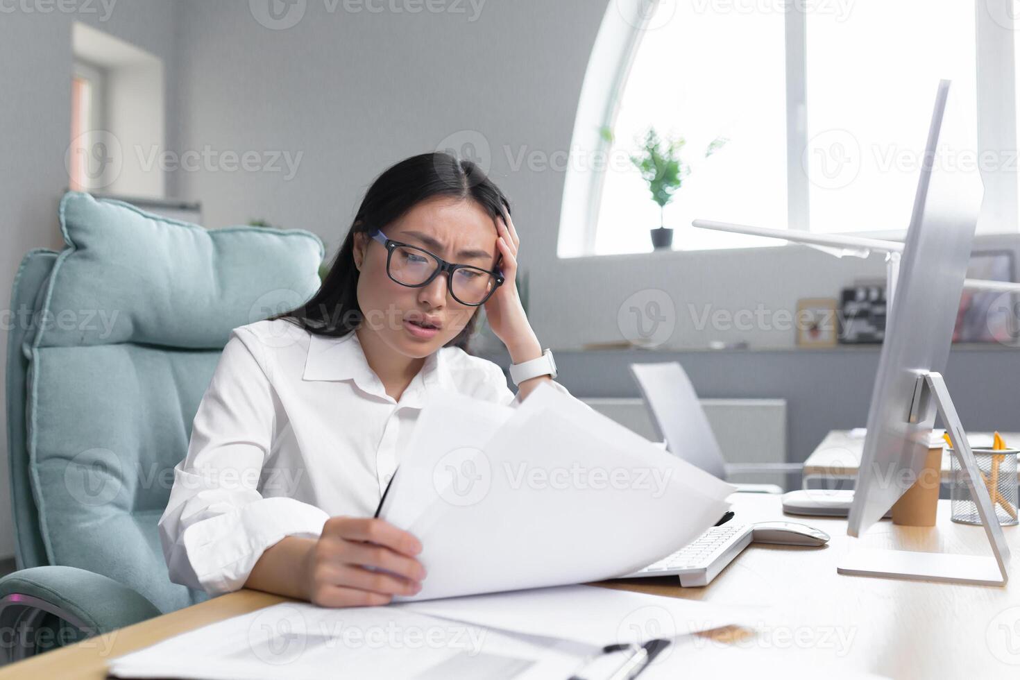 Deadlines are burning. Young beautiful Asian businesswoman is worried, sits in the office at the desk, holds documents in her hands. Shows them, holds her head with her hands, does not know what to do. photo