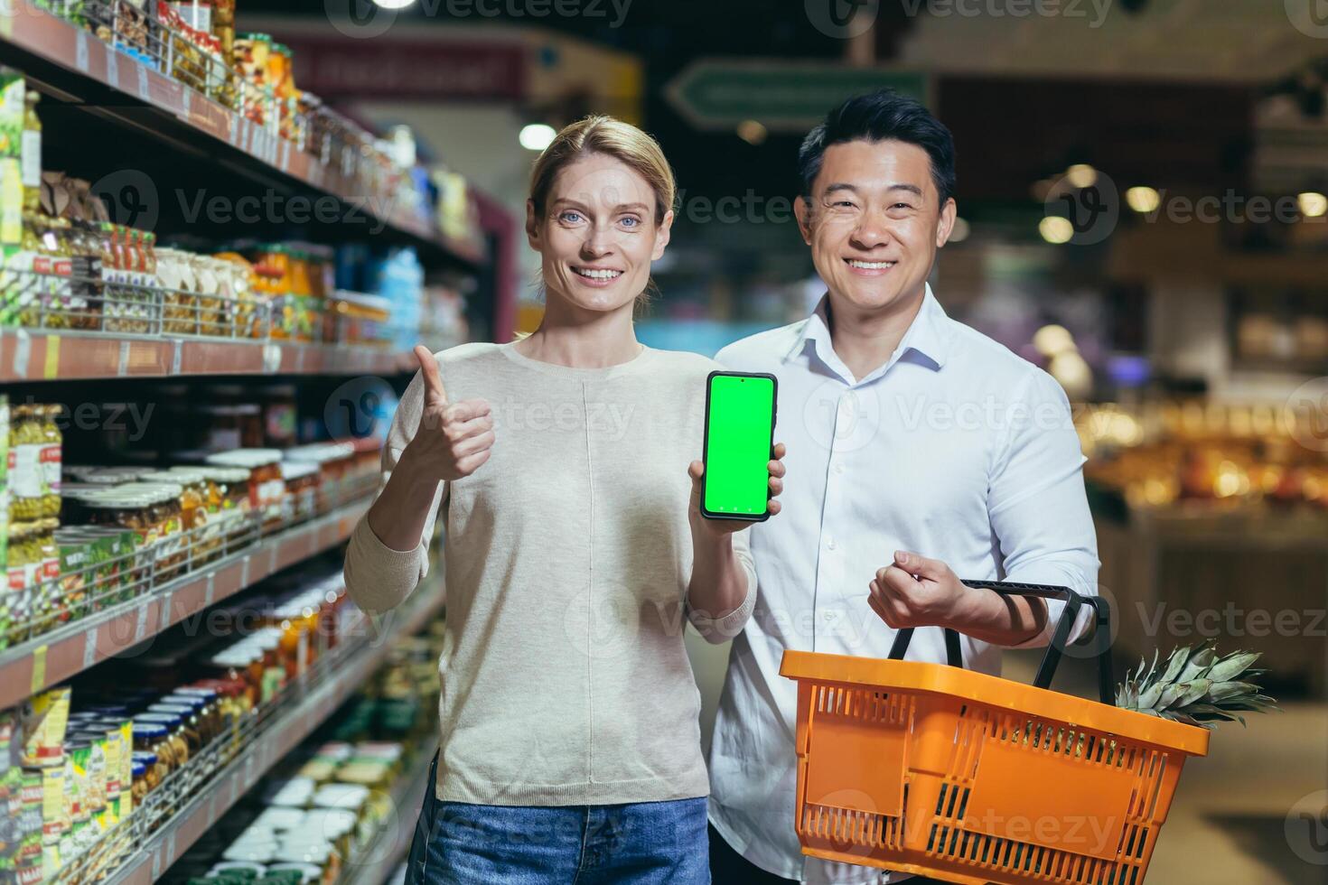 Portrait of a happy Asian couple of consumers, shoppers in a supermarket or grocery store, looking at the camera, smiling. are holding a phone and showing a green chromakey screen photo