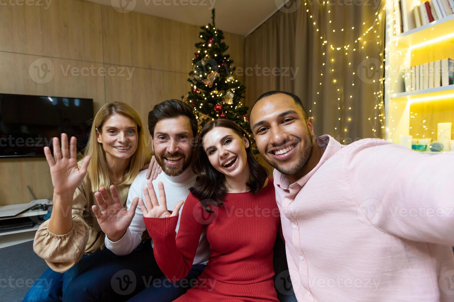 Diverse friends celebrating christmas and new year together, group of people having dinner at home sitting on sofa near Christmas tree, taking selfie photo and talking with friends on call using phone.