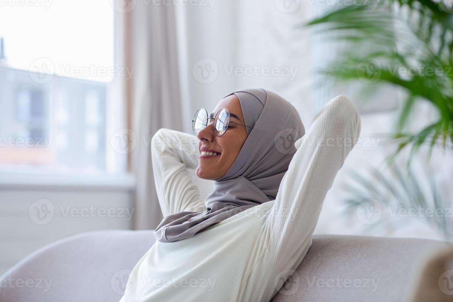 Close-up photo. Smiling Muslim young woman in hijab and glasses sitting on sofa at home, hands behind head and resting, eyes closed, smiling, meditating, dreaming, relaxing. photo
