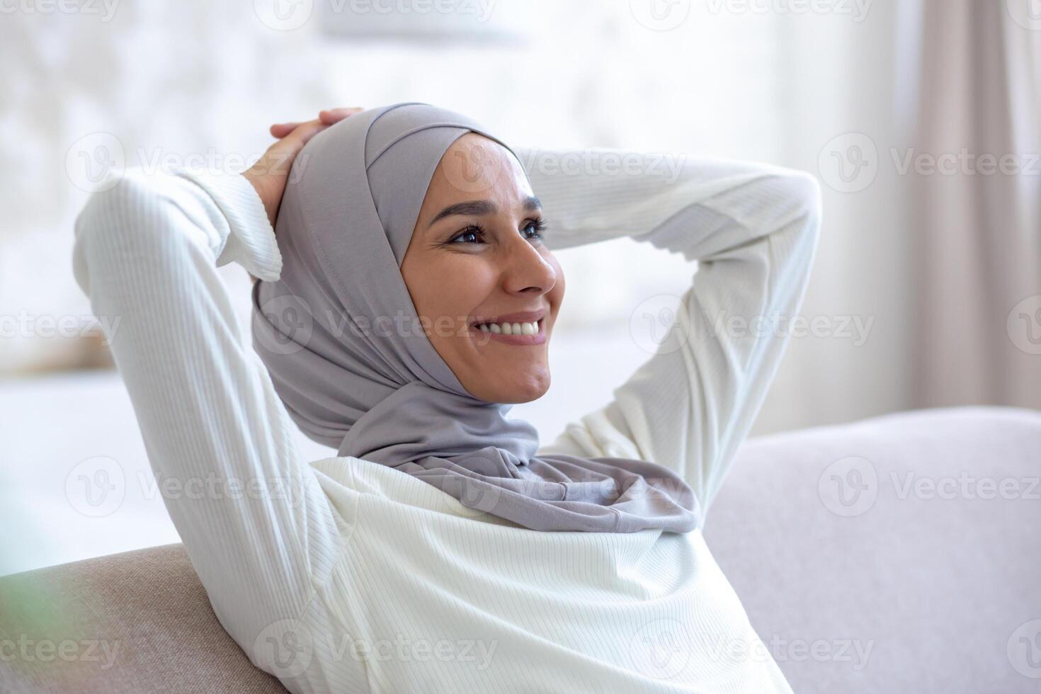 Close-up photo. Smiling arab young business woman in hijab sitting on sofa at home with hands behind head and resting, smiling and satisfied with successful work done and finished. photo