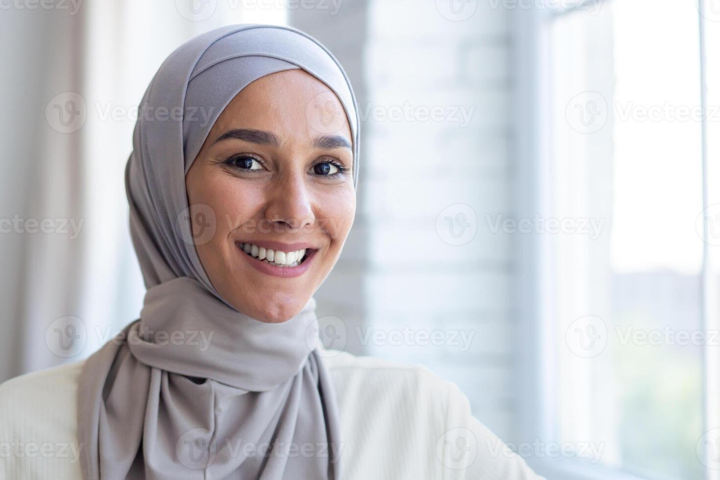 Close-up portrait of young beautiful arab woman, muslim woman in hijab smiling and looking at camera, standing near window at home. photo