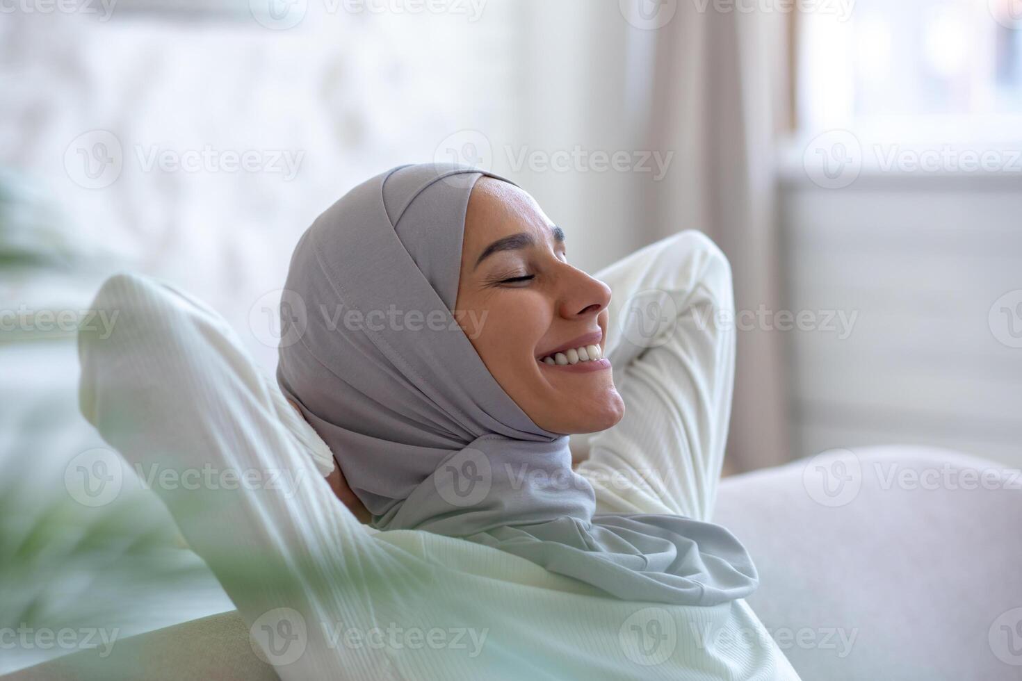 Close-up photo. Smiling Muslim young woman in hijab sitting on sofa at home and resting, eyes closed, smiling, meditating, dreaming, relaxing, worrying about her mental and psychological health. photo